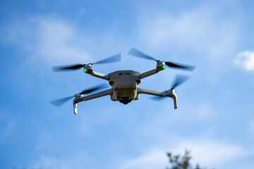 Close-up of a quadcopter drone flying in the sky on a bright day. The white drone is hovering mid-air with its propellers spinning, against a clear blue sky background. © daily_creativity