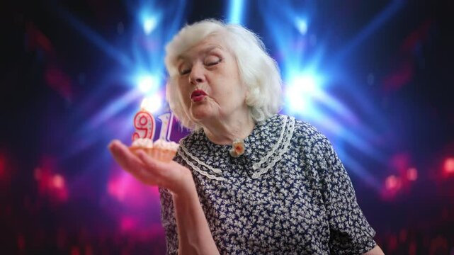 Delighted senior woman blowing out candles on birthday cake in a concert hall