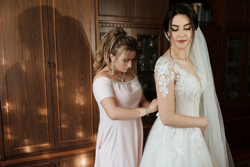 A bride is getting ready for her wedding, with her bridesmaid helping her adjust her dress