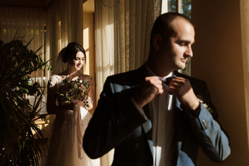 A man is getting ready to tie his tie while a woman stands behind him. The scene is set in a room with a window, and the man is wearing a suit. The woman is holding a bouquet of flowers