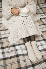 Woman in a knitted sweater sitting on a cozy plaid blanket, holding a cup of hot chocolate or cocoa drink with marshmallows. Hygge concept