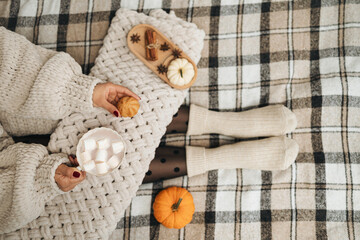 Woman in a knitted sweater sitting on a cozy plaid blanket, holding a cup of hot chocolate or cocoa drink with marshmallows. Hygge concept