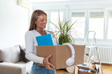 Happy woman carrying her belongings while relocating into new apartment. Young woman holding a box with plants and files in new home.