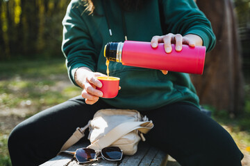 A woman is meticulously pouring steaming, aromatic coffee from her thermos into a charming cup, savoring a peaceful moment of relaxation outdoors amidst the beautiful trees and soft grass