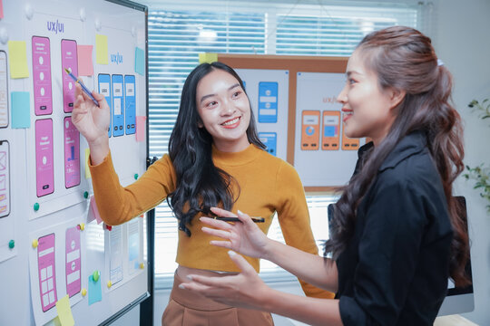 Two asian ui/ux designers are discussing mobile app interface wireframe design on whiteboard, working together in the office