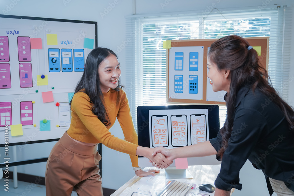 Wall mural two young asian women, both ui/ux designers, happily collaborating on a mobile app interface in an o