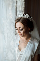A woman in a white dress is wearing a tiara and veil. She is standing in front of a window, looking...