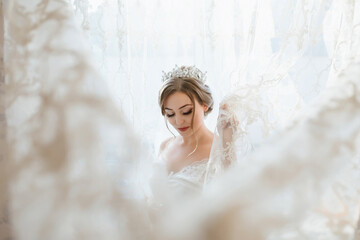 A woman in a white dress is standing in front of a curtain. She is wearing a tiara and has her hair in a bun
