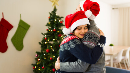 Young couple wearing festive sweaters and santa hats hugging each other near a decorated christmas tree in their home