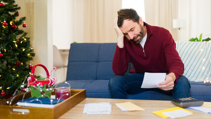 Worried man reviewing his home finances during christmas time while holding a document and sitting on a couch
