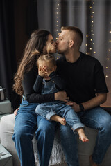 Naklejka premium Caucasian family capturing intimate moment with young parents kissing while child playfully covers eyes. Parents wear casual clothing sitting in cozy home setting.