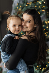 Young Caucasian mother holding daughter in front of Christmas tree. Both smiling, wearing black tops and denim. Festive, warm atmosphere, showcasing family bond and joy during holiday season.