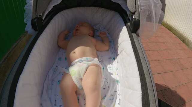 A infant lying on its back in a pram cradle. The toddler The pram is driving in the backyard, green fence and red tiles are visible