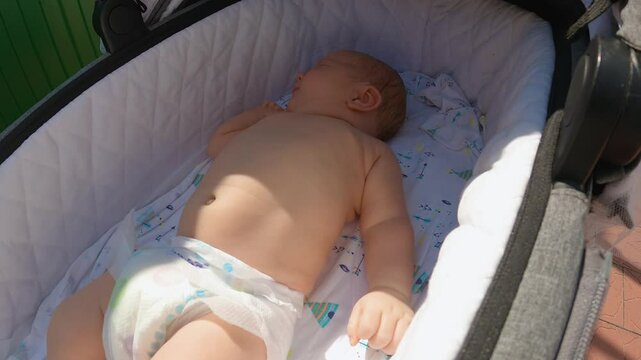 An infant is lying in a pram cradle with one nappy on. Close-up view. A walk with the baby in the fresh air