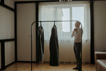 A man is standing in front of a clothes rack with a dress hanging from it. He is adjusting his tie and looking at the dress. The room is dimly lit, and the dress is black