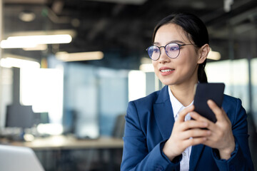 Asian businesswoman in professional attire holding smartphone in modern office setting. Confident expression conveys success and productivity in business environment. Represents technology