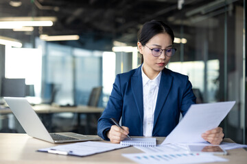 Asian businesswoman concentrating on document review at office desk. Equipped with laptop and...