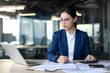 Asian businesswoman working diligently in office setting, focused on laptop and paperwork, using calculator. Represents dedication, focus, and professionalism in workplace environment.
