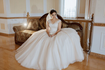 A woman in a white dress sits on a couch. She is wearing a tiara and a veil