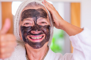 Cheerful beautiful senior woman in bathrobe applying a detox facial charcoal mask homemade looking...