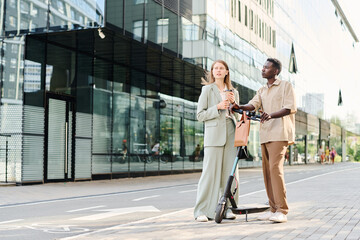 Wide shot of Caucasian woman holding cup of coffee and her Black male colleague with e-scooter chatting while standing outdoors