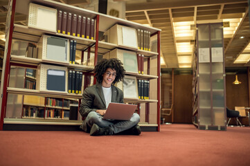 Young curly-haired man sitting on the floor in the library with a laptop in hands