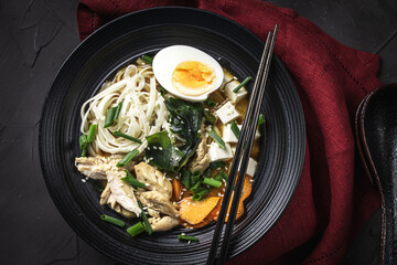 Japanese miso soup with udon noodles, tofu cheese, chicken, green onions and sesame seeds in a black bowl with chopsticks and a burgundy napkin on a dark background. Top view. Copyspace