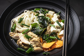 Japanese Miso Soup with udon noodles, tofu cheese, chicken, green onions and sesame seeds in a black bowl with chopsticks on a dark background. Close-up. Top view. Copyspace
