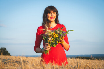 Glückliche junge und attraktive Frau steht mit einem Blumenstrauss im Kornfeld.