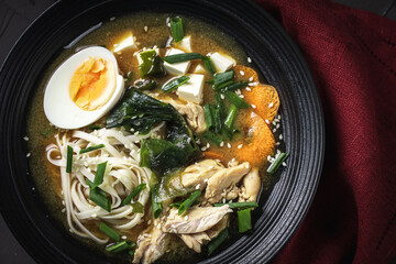 Japanese miso soup with udon noodles, tofu cheese, chicken, green onions and sesame seeds in a black bowl with chopsticks and a burgundy napkin on a dark background. Top view. Copyspace