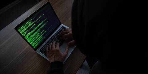 Man in a dark hoodie types on a laptop displaying green code on the screen. The image emphasizes themes of hacking, programming, cybersecurity, or tech-related activities in a dimly lit setting.