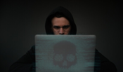 Young hacker man in a hoodie and glasses works on a laptop displaying a skull image made from digital code. The image conveys hacking, cybersecurity threats, malware, or digital danger themes.
