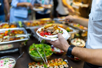 A person serves themselves a hearty meal at a bustling buffet during an evening gathering filled...