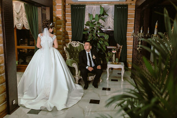 A bride and groom are standing in front of a green chair. The bride is wearing a white dress and the groom is wearing a black suit. Scene is romantic and happy