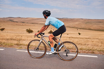 Fototapeta premium A cyclist rides a road bike on a paved road through a beautiful rural landscape. The rider is wearing a helmet and a blue cycling jersey. The scenery is dominated by brown fields and a clear blue sky.