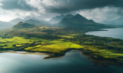 Obraz premium Aerial view of tranquil shoreline, green fields, and majestic mountains under a cloudy sky