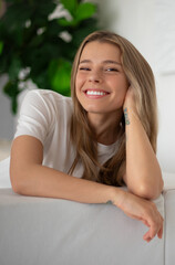 Naklejka premium Gorgeous woman with long blonde hair smiles brightly while leaning on couch, wearing white t-shirt. The relaxed, cozy indoor setting features greenery and soft lighting, evoking warmth and happiness