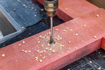 A person is using a drill to make a hole in a piece of wood. The drill is making a hole in the wood and wood shavings are flying around. Construction of a house, construction of a foundation.