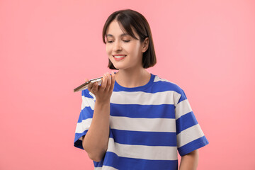Beautiful woman recording voice message via smartphone on pink background