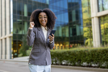 African American business woman standing outside modern office building holding phone looking excited and confident. Expression of success, professional achievement, and positive energy in business