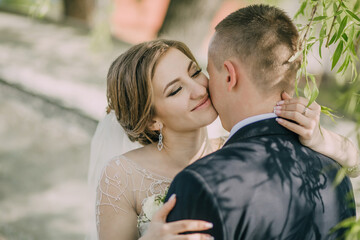 A bride and groom are hugging each other in the shade. The bride is wearing a white dress and the groom is wearing a blue suit