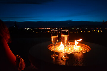 A dimly lit woman in evening wear alongside two glasses of champagne and fire pit on a patio overlooking city lights at blue hour evening twilight.