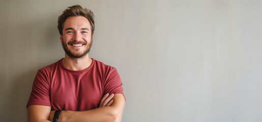 A smiling young man with a beard and crossed arms stands confidently against a neutral background, exuding warmth and approachability