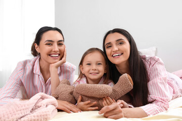 Fototapeta premium Young lesbian couple with adopted little girl and toy lying in bedroom