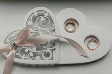 A white heart-shaped box with a pink ribbon and two gold rings inside. The box is decorated with a lace design and the rings are placed on top of each other. Concept of love and commitment