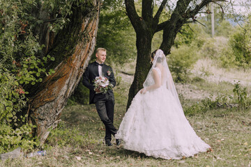 A bride and groom stand in front of a tree, with the bride wearing a white dress and the groom wearing a suit