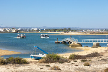A tranquil scene in Fuseta, Algarve, showing boats navigating the calm lagoon waters near a small...
