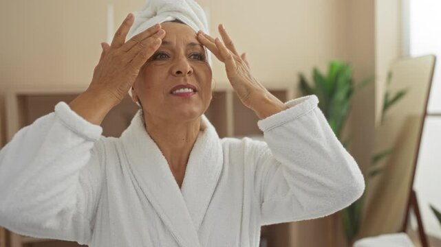 Woman enjoying a spa day in a wellness center, wearing a white bathrobe and towel, touching her face for skincare routine, surrounded by indoor plants and spa decor.