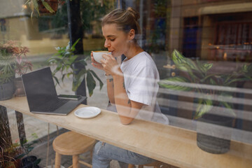 A young woman sips coffee, engaged in work at a cafe with plants nearby, showcasing a serene workspace.
