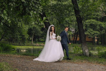 A bride and groom are walking through a forest, holding hands and smiling. The bride is wearing a white dress and a veil, while the groom is wearing a suit. The scene is peaceful and romantic